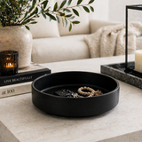 Matte black round decorative tray used as an entryway organizer holding keys and accessories on a modern console table with candle, books, and greenery