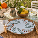 Ceramic dinner plate with green botanical leaf and berry pattern on outdoor table setting with lemons, flowers, and rustic decor in natural sunlight