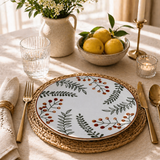 Elegant table setting with botanical ceramic plate featuring green leaves and red berries, styled with lemons, candlelight, glassware, and warm natural lighting