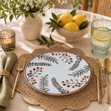 Botanical ceramic plate with leaf and berry pattern on woven rattan placemat, styled with lemons, glassware, candle, and floral arrangement in warm sunlight