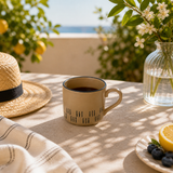 Beige mug with black patterns on a table with a hat, flowers, and fruit in the background.