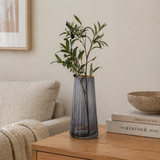 Minimalist living room styling with a smoky glass vase holding olive branches on a wooden side table beside neutral decor and stacked books