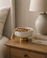 Neutral bedside table styling with a ceramic bowl filled with wooden beads, textured lamp, and stacked books, creating a warm modern minimalist home decor look