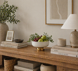 Neutral console table styled with a ceramic fruit bowl filled with apples and grapes, layered books, table lamp, and greenery, creating a warm modern home decor vignette
