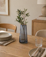 Modern dining table styling with a smoky glass vase holding olive branches surrounded by neutral tableware, wood furniture, and soft minimalist decor