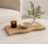 Organic shaped wooden textured decorative tray styled with candle, bowl, and glass vase on a modern coffee table in a soft minimalist living room setting