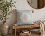 Pastel geometric throw pillow with pink tassels styled on a woven bench in a cozy entryway with neutral decor and greenery