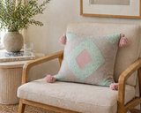 Pastel geometric throw pillow with pink tassels styled on a neutral accent chair beside a side table with decor and greenery