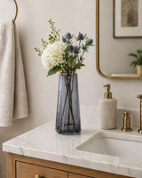 Smoky glass vase with white hydrangea and blue thistle flowers styled on a marble bathroom countertop with brass fixtures and soft neutral decor