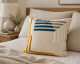 Textured geometric throw pillow with blue, gray, and mustard accents on a neutral bed in a cozy modern bedroom setting