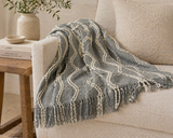 Textured gray throw blanket with tassels draped over a neutral sofa in a cozy modern living room setting
