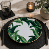 Tropical leaf porcelain dinner plate with gold rim styled on a modern black table setting with dark placemat, candle, glassware, and greenery