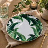 Tropical leaf porcelain dinner plate with gold rim styled on a warm natural table setting with linen napkin, candle, greenery, and textured glassware